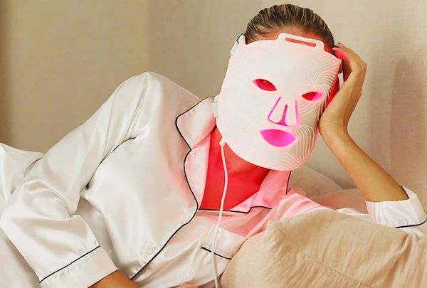 A woman relaxing in bed wearing elegant white silk pajamas with black piping. She is wearing a flexible white LED facial mask that is emitting a bright red light. She is resting her head comfortably on her hand against soft pillows, enjoying an at-home light therapy session.
