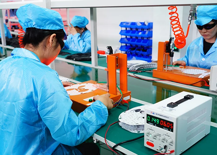 Wakelife factory production line showing technicians in blue cleanroom suits and safety goggles working at a green workbench. They are using an orange testing fixture and a DC power supply to test and assemble the white internal LED light panels for facial masks.